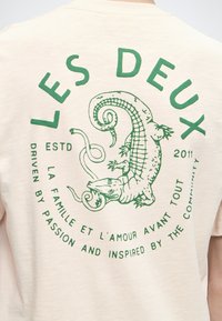Graphique verte d'un crocodile enroulé avec le texte « LES DEUX » et des phrases en français et en anglais au dos d'un t-shirt beige clair.