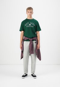 Grønn bomulls T-skjorte med "GAP 1986 ATHLETICS" trykk, grå joggebukser og en rød rutete flanell skjorte knyttet rundt livet, svarte sneakers.