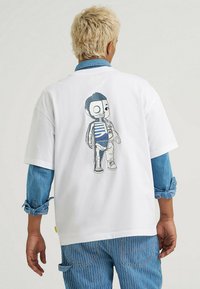 Biała bawełniana koszulka z nadrukiem postaci szkieletu w stylu kreskówkowym na plecach, w paskowanej koszuli i niebieskich dżinsach, z podwiniętymi rękawami.