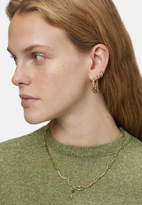 Guldtonede metal øreringe med unikke organiske former og en tekstureret overflade, parret med en guldkæde necklace, båret over en grøn sweater.