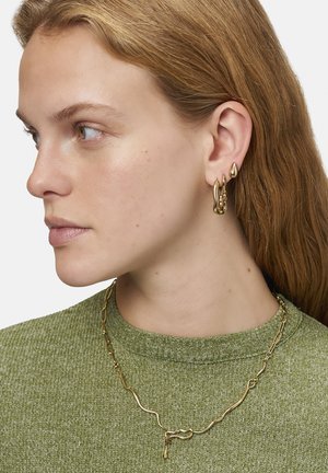Guldtonede metal øreringe med unikke organiske former og en tekstureret overflade, parret med en guldkæde necklace, båret over en grøn sweater.