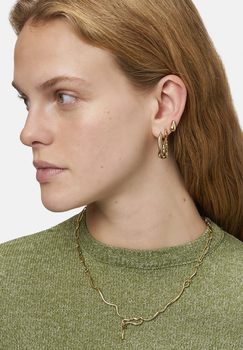 Guldtonede metal øreringe med unikke organiske former og en tekstureret overflade, parret med en guldkæde necklace, båret over en grøn sweater.