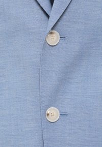 Šviesiai mėlynas lino švarkas su negilia apykakle, dviem priekiniais mygtukais ir subtilia tekstūra. Mygtukai yra kreminės spalvos su keturiomis skylutėmis.