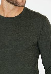 Musta vihreä pitkähihainen paita pehmeästä, tekstuuriltaan mielenkiintoisesta kankaasta, jossa on pyöreä kaula-aukko ja hienovarainen vaakasuora kuvio.