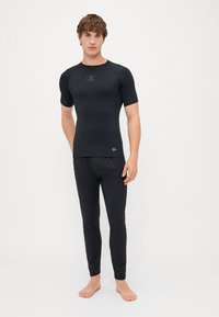 Haut de sport noir à manches courtes avec logo, associé à un legging noir ajusté. Matériau lisse et extensible, conçu pour le confort et la mobilité.