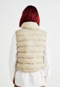 Chaleco acolchado beige con cuello alto, con acolchado horizontal y una textura suave, llevado sobre una camisa blanca y pantalones marrones.