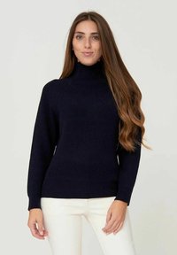 Maglione a collo alto a coste blu navy con maniche lunghe. Presenta un design aderente, un tessuto testurizzato e una finitura liscia. Pantaloni bianchi indossati sotto.