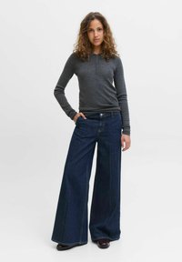 Žena s kudrnatými vlasy stojí s jednou rukou v kapse, oblečená v tmavě šedém tričku s dlouhým rukávem a širokých tmavě modrých džínách na bílém pozadí.