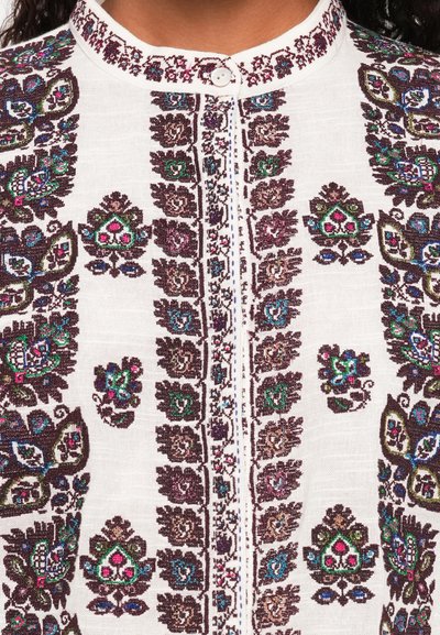 Fehér hímzett blúz, amely bonyolult virágmintákkal díszített sötétbarna, zöld és kék színekben. Gombos záródás a nyakrésznél.