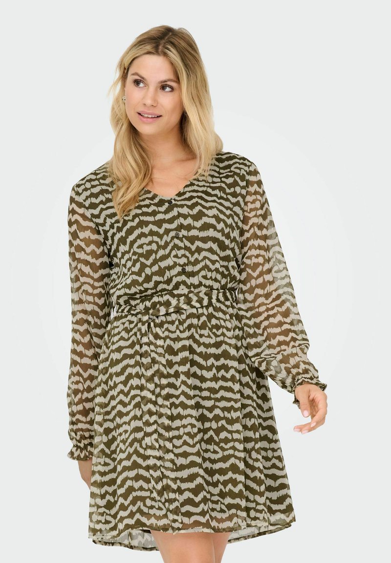 Vrouw met lang blond haar, gekleed in een olijfgroene en beige gestreepte jurk tot op de knie met lange mouwen en een riem, kijkt opzij.
