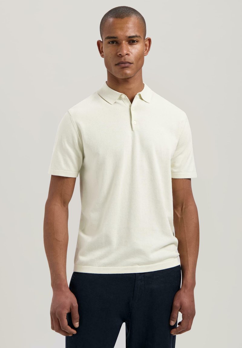 Lys kremfarget polo-skjorte laget av myk stoff, med ribbestrikket krage, korte ermer og en tettsittende design. Kombinert med mørke bukser.