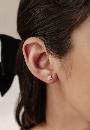 Silberne Steckerohrringe mit floralem Design und drei funkelnden Edelsteinen. Nahaufnahme eines Ohrs gegen dunkles Haar.