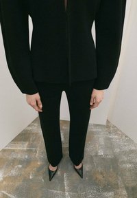 Top negru mulat cu mâneci lungi și fermoar frontal, asortat cu pantaloni negri cu croială dreaptă. Purtați cu pantofi negri cu toc ascuțit, pe un podea gri texturată.