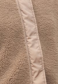 Chaqueta de forro polar en marrón claro con una superficie texturizada y esponjosa, y un panel suave en beige claro que contrasta a lo largo de la cremallera.