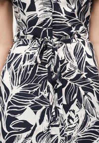 Κοντινό πλάνο ενός φορέματος με τροπικό μοτίβο σε ναυτικό μπλε και λευκό, με κουμπιά στο μπροστινό μέρος και δεμένη υφασμάτινη ζώνη στη μέση.