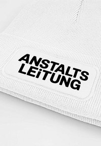White knitted beanie featuring a rectangular patch with black text reading "ANSTALTS LEITUNG." Text is bold and prominently displayed.