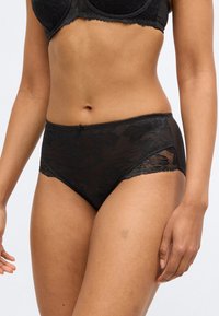 Črne hlačke z visokim pasom, okrašene z cvetličnim čipkastim prekrivanjem in prosojnimi stranicami. Gladek teksturni material z valovitimi robovi in elastičnim pasom.