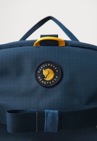 Mornarsko plavi ruksak od teksturirane tkanine, s žutom drškom, tamnoplavim detaljima i kružnim logotipom sa stiliziranim dizajnom životinje.
