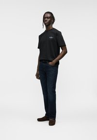 Μαύρο βαμβακερό t-shirt με κοντά μανίκια, με μικρό κεντημένο λογότυπο. Φορεμένο με σκούρο μπλε τζιν και καφέ παπούτσια τύπου slip-on.