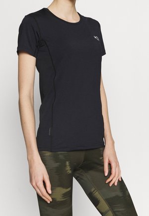 Persoon die een nauwsluitend zwart sport-T-shirt met korte mouwen en groene camouflage legging draagt, staand tegen een effen achtergrond.