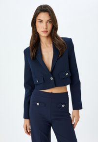 Lyhennetty tummansininen blazer syvällä V-kaula-aukolla, hopeisilla nappikoristeilla ja kahdella etutaskulla. Yhdistetty samanvärisiin muotonsa mukaisiin tummansinisiin housuihin. Sujuva pinta.