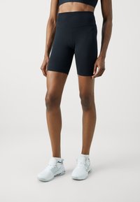 Czarne rowerowe szorty z wysokim stanem, wykonane z elastycznego materiału, o gładkiej teksturze. Noszone z białymi sportowymi butami na jasnym tle.