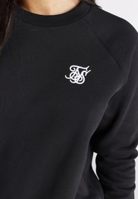 Zwarte trui met een geribde ronde hals en raglanmouwen. Voorzien van een wit geborduurd logo op de linkerborst. Glad stoffen oppervlak.