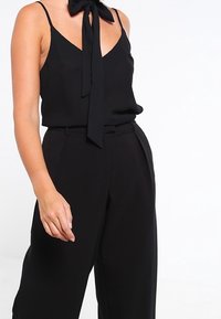 Black sleeveless top with a bow tie detail at the neckline, paired with high-waisted black pants featuring a tailored design and wide legs.