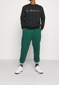 Černý pruhovaný svetr s bílým logem, kombinovaný se zelenými tepláky s bočními kapsami a žebrovanými manžetami na kotnících, obutý do bílých tenisek.