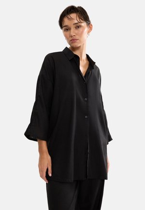 Kvinde med kort hår iført en løs sort skjorte med knapper og brede ærmer samt matchende sorte bukser mod en ensfarvet baggrund.