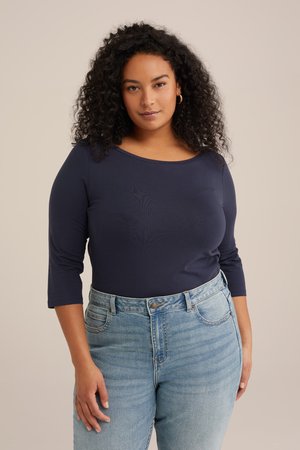 Femeie cu păr creț, purtând un top cu mâneci lungi, de culoare albastru închis, și blugi cu talie înaltă, de culoare albastru deschis, stând pe un fundal simplu bej.