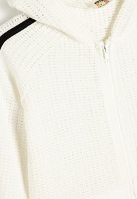 Haut à capuche zippé blanc en tissu texturé et respirant, avec une bande d'accent noire et un cordon de serrage.