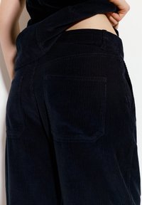 Temno modri kordurojski hlače z vertikalnim ribanjem, opremljene z dvema zadnjima žepoma in brezšivnim pasom, ki poudarja njihovo teksturirano blago.