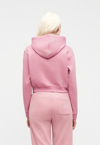 Γυναίκα με μακριά ξανθά μαλλιά που φοράει ροζ κοντή φούτερ μπλούζα με κουκούλα και ροζ παντελόνι φόρμας με λογότυπο σε πίσω τσέπη, γυρισμένη πλάτη σε λευκό φόντο.