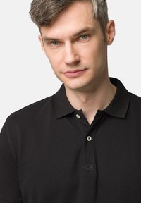 Czarna koszulka polo z krótkim rękawem, z zapięciem na dwa guziki i kontrastującymi jasnymi guzikami. Tkanina ma teksturowany splot.