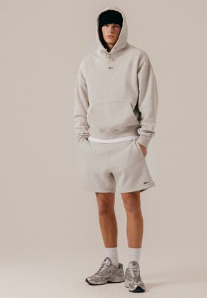 Ung mand iført lysegrå hættetrøje og matchende shorts med hvide sokker, sølvfarvede sneakers og en sort beanie, stående med hænderne i lommerne.