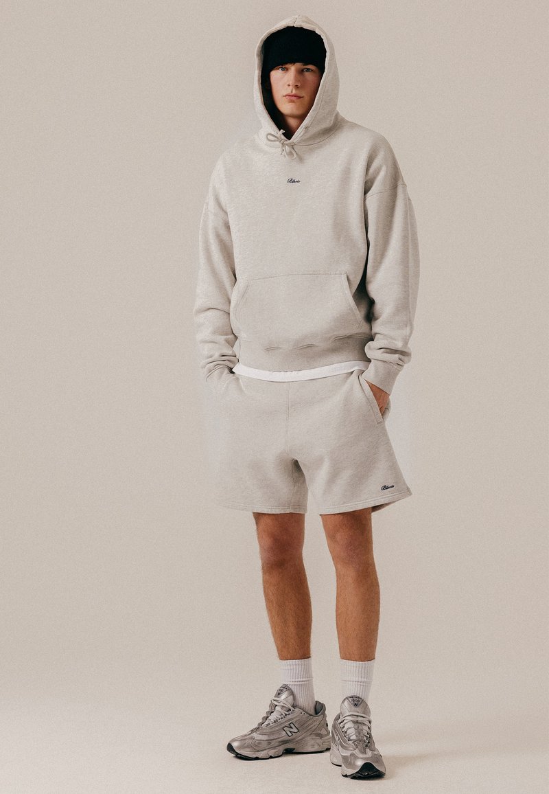 Jeune homme portant un sweat à capuche gris clair et un short assorti, des chaussettes blanches, des baskets argentées et un bonnet noir, debout les mains dans les poches.