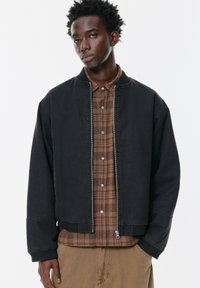 Crna bomber jakna s rebrastim ovratnikom, zatvaranjem na patentni zatvarač i spuštenim ramenima, složena preko smeđeg kariranog košulje s kopčama na gumb.