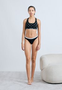 Μαύρο αθλητικό σουτιέν με λευκό σχέδιο αστεριού και λωρίδα με λογότυπο, σε συνδυασμό με μαύρο κάτω μέρος μπικίνι με λευκή μέση, πάνω σε ανοιχτό χαλί.