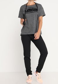 Šedé tričko s krátkým rukávem a černým logem, doplněné černými sportovními kalhotami a světle růžovými teniskami; skákací guma přehozená přes krk.