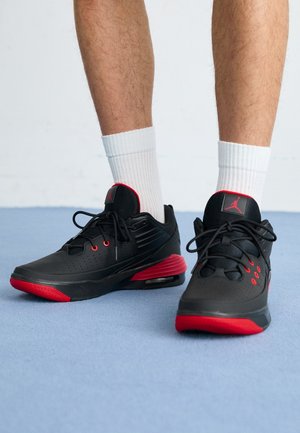 Personne portant des chaussures de sport noires et rouges avec des chaussettes blanches, debout sur un sol bleu clair.