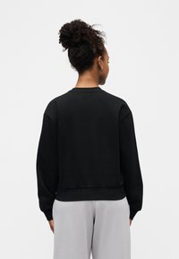 Femme aux cheveux bouclés attachés en chignon, portant un haut noir à manches longues et un pantalon gris clair, debout de dos face à la caméra.