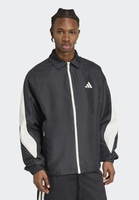 Sort og hvid atletisk jakke med krave, frontlynlås og elastiske manchetter. Bemærkelsesværdigt Adidas-logo på venstre bryst.