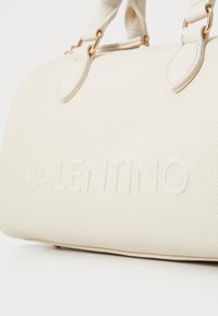Sac en cuir beige à texture granulée avec logo "VALENTINO" en relief, doté de deux poignées supérieures et d'accents en métal doré.