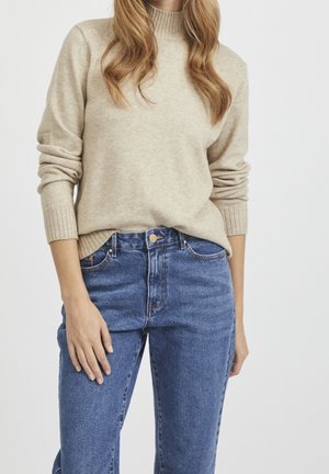 Frau trägt einen beigen, gerippten Pullover mit langen Ärmeln und blaue, hoch taillierte Jeans, steht vor einem einfarbigen Hintergrund.