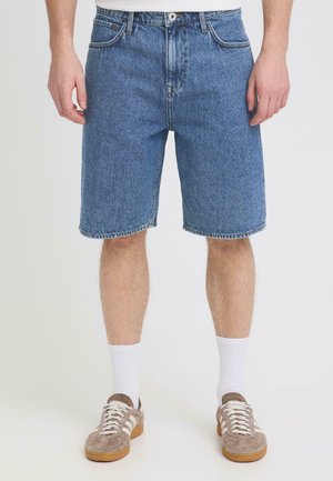 Mężczyzna w niebieskich dżinsowych szortach o długości do połowy uda, białych skarpetkach do kostki i beżowych trampkach, stojący na jednolitym jasnym tle.