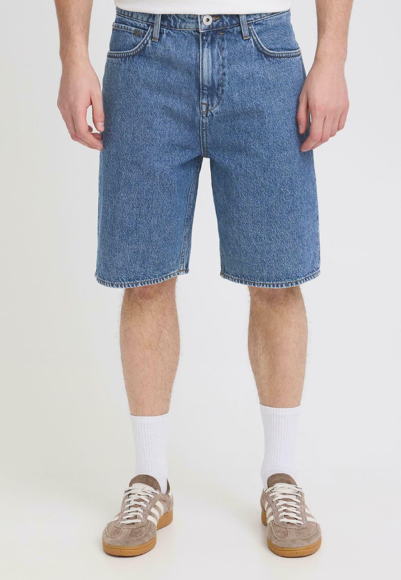 Homme portant un short en jean bleu mi-long, des chaussettes blanches hautes et des baskets beiges, debout devant un fond clair uni.