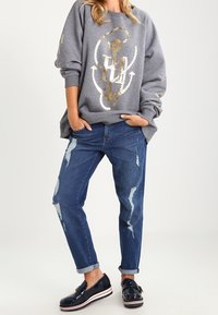 Harmaa huppari, jossa on kultainen graafinen printti, löysä istuvuus; yhdistettyinä repaleisiin sinisiin farkkuihin ja kiiltäviin mustiin kenkiin, joissa on valkoinen pohja. Rento asu.