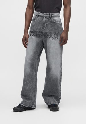 Personne portant un jean gris à jambes larges avec un texte gothique noir imprimé sur le haut des cuisses et des baskets noires.