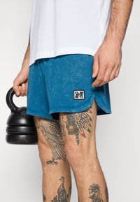 Plave pamučne šorts hlače sa izblijedjelom teksturom, s crnim Nike logom pri rubu. U lijevoj ruci drži girju.